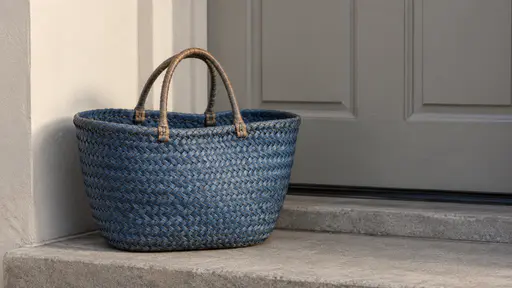 A blue woven market basket with worn handles rests empty on a plain doorstep beside the door in morning light.