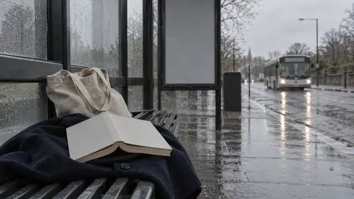 A paperback with blank pages lies open on a folded navy coat on the bench of a bus shelter with a canvas tote bag beside it and a bus approaching down a rain-wet road in flat grey light.