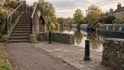 The landing at the end of a canal footbridge in warm late-afternoon light has a small paved widening with a low stone wall and two iron bollards while the footbridge steps and towpath stay clear and narrowboats are moored in the canal beyond.