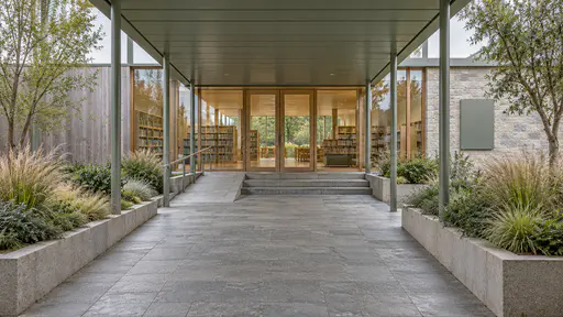 A wide paved approach runs under a long sage green canopy on slim steel posts to the timber-framed glass entrance of a small library with grass planters on either side in soft overcast light.