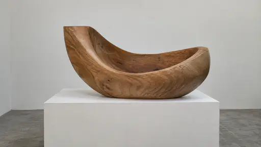 A curved carved wooden form with one deep smooth hollow rests on a pale plinth against a plain wall in even gallery light.