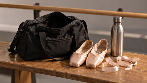 On a studio bench in warm side light a black dance bag stands open with a pair of pink ballet slippers, two satin ribbons and a metal water bottle laid out beside it for rehearsal with a bare wooden barre blurred behind.