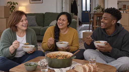 A woman with an auburn bob in a sage-green cardigan, a woman with a long braid in a mustard jumper and a young man in a charcoal hoodie sit on a living-room floor around a low table sharing a one-pot meal with unopened boxes behind them in soft lamp light.