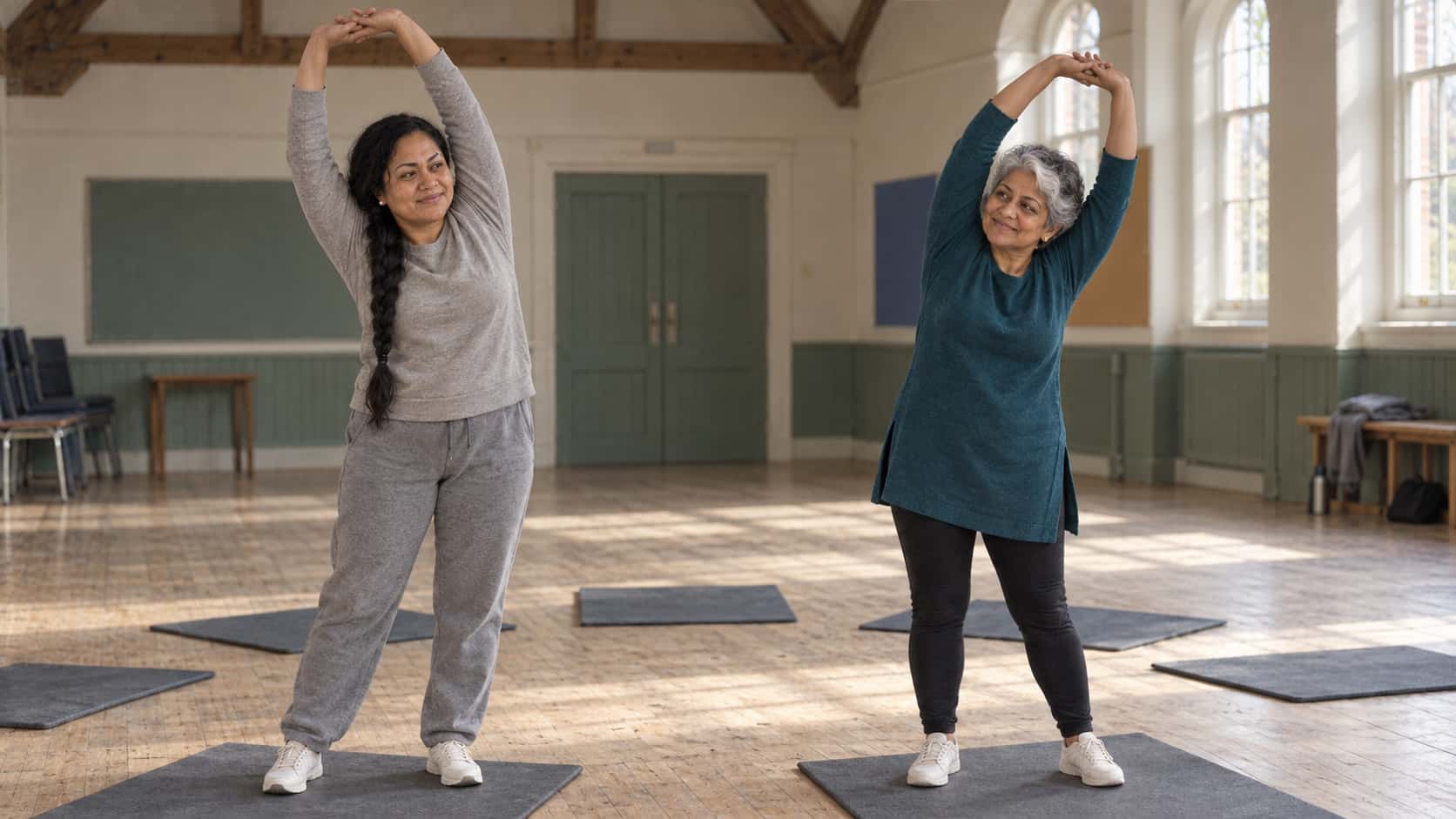 Two women, one with a long black braid in a grey tracksuit and one silver-haired in a teal tunic, stretch with raised arms on foam mats in a circle with empty mats between them in a timber-beamed hall in low morning light.