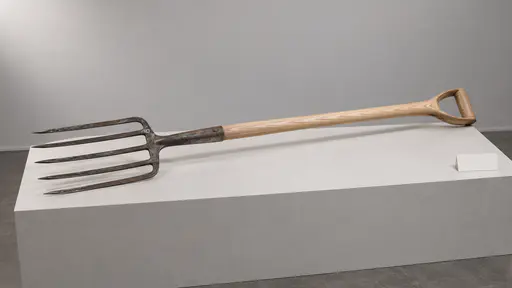 A garden fork with an old steel head and a pale new ash handle lies diagonally on a long museum plinth with a blank label card at one end.