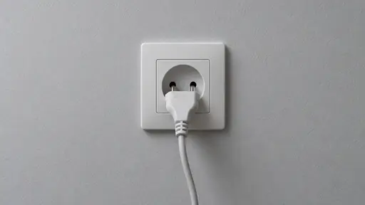 On a plain painted wall in even frontal light a white two-pin plug is seated exactly halfway into a white wall socket with its pale grey cable curving down out of the frame bottom and both plug and socket fully in view seen straight on.