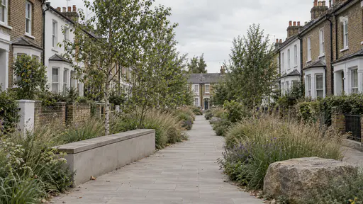 A residential lane in soft overcast light widens into a planted verge of grasses and small trees with a low wall to sit on and a clear paved walking strip running straight through the middle of the planting between terraced houses.