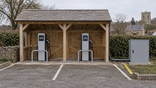 An open-fronted timber charging shelter with a pitched roof covers two plain charge posts with hung cables at the edge of a village car park with a marked duct leading to a grey cabinet beside it under flat overcast light.