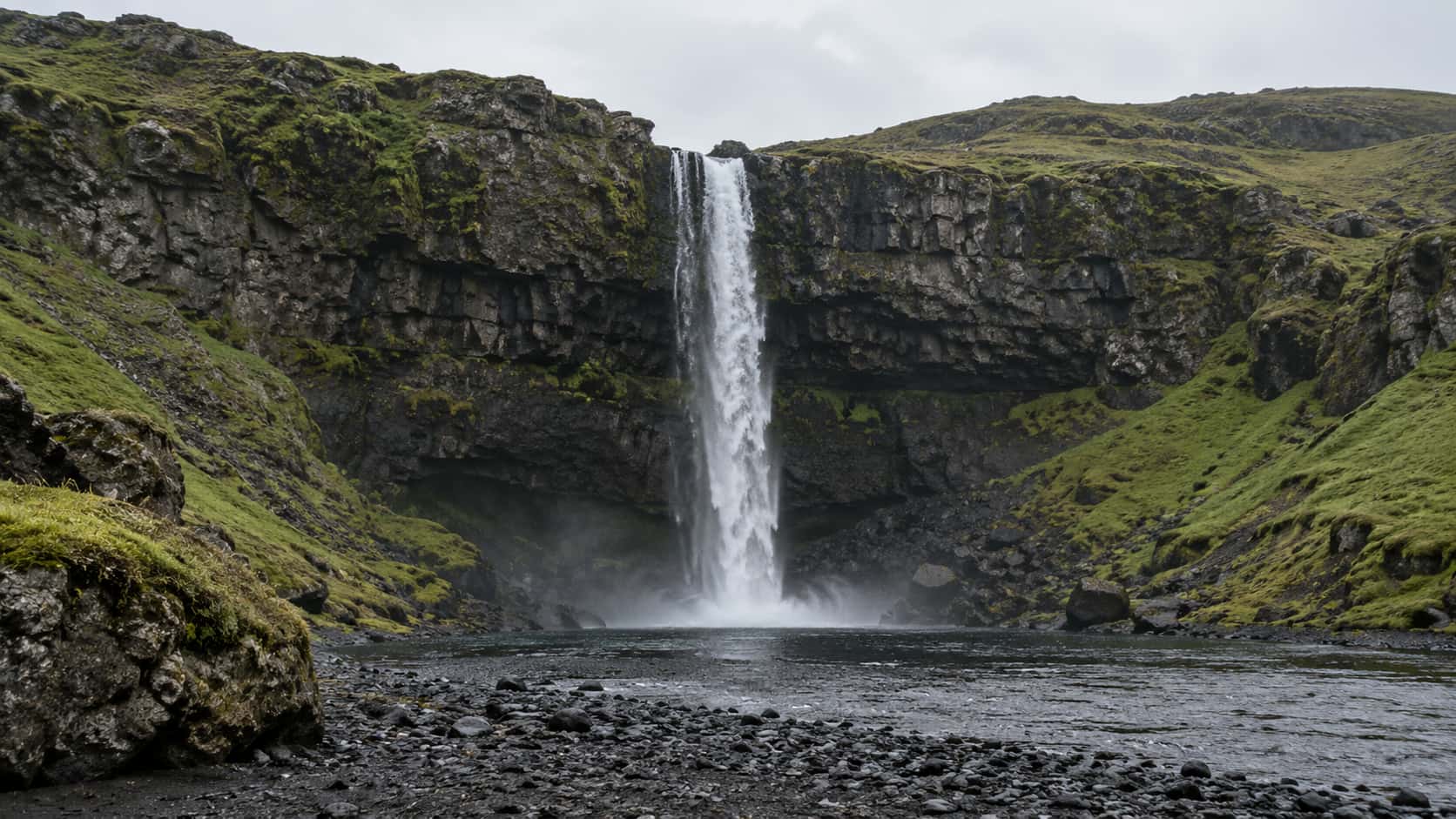 A tall narrow waterfall drops in one unbroken white ribbon over a dark cliff ledge into a black pebble shore pool, green moss on the cliff sides.