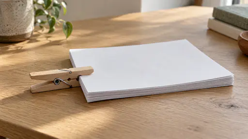 A plain wooden spring clothes peg with two smooth jaws is clamped over the left edge of a neat stack of about ten blank white sheets with its upper jaw resting on the top sheet and its lower jaw under the bottom sheet, the sheets visibly squeezed between the two jaws, the stack lying flat on a plain light oak desk in soft side daylight seen from a slight high angle.