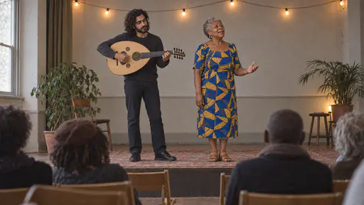 A slim bearded musician plays an oud on a low stage while an older woman with short silver curls in a blue and yellow wax-print dress stands beside him singing a verse with her head lifted and one hand open at her side.