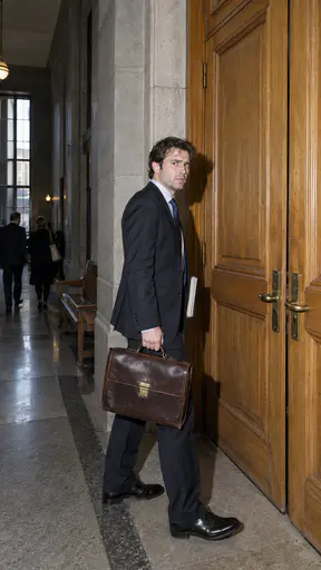 A fictional adult man lawyer in a dark suit holds a leather briefcase and pauses before tall wooden courtroom doors in a stone corridor in a hard direct flash photograph.