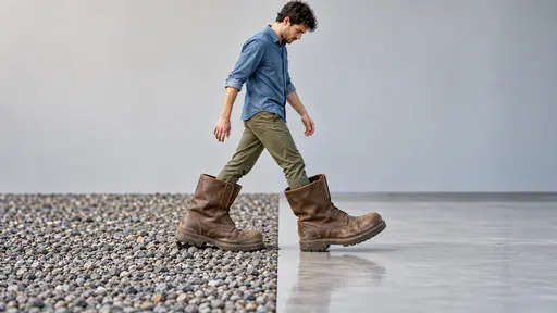 Walking in someone else's shoes: a small adult walks mid-stride inside a visibly oversized pair of worn work boots, looking down carefully, the left boot pressing coarse gravel while the right boot steps over smooth office flooring in clear daylight.