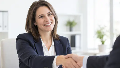 A fictional adult woman with straight hair in a navy blazer and white shirt smiles confidently and offers a handshake to a colleague at the edge of the frame in a bright white office.