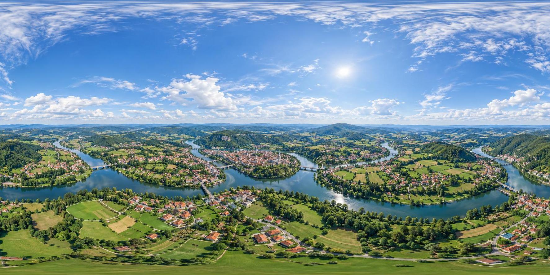 A full 360 degree equirectangular aerial panorama from high above a green river valley town with red roofed houses, a winding river, patchwork fields and forested hills with sky overhead.