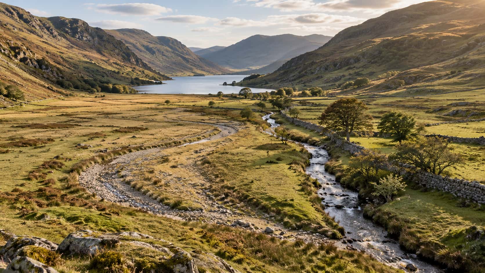 The dried curve of an old meander lies across a valley floor as a pale grass-filled scar with a stony gravel bed while the living stream winds narrow along a drystone wall beside it in raking late light.