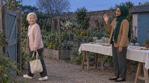 A very small elderly woman with a low silver bun in a dusty-pink cardigan walks out through a garden gate carrying a cloth tote of small pots and looks back smiling while a young woman in a green hijab waves beside the bare trestle table at blue dusk.