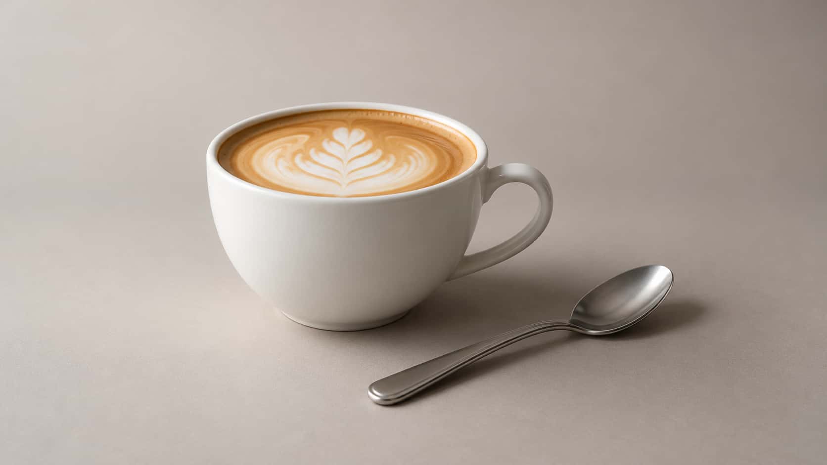 One plain latte cup rests on a plain surface in even daylight beside one clean unused plain spoon lying flat.