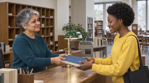 A petite woman with short silver hair in a teal sweater holds a plain blue hardback book out across a wooden library counter to a smiling young woman with a short natural afro in a yellow sweatshirt reaching for it with both hands.