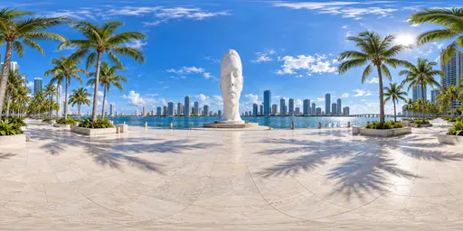 A panoramic view of a bayfront plaza with palms, skyline, water and a large white abstract head sculpture.