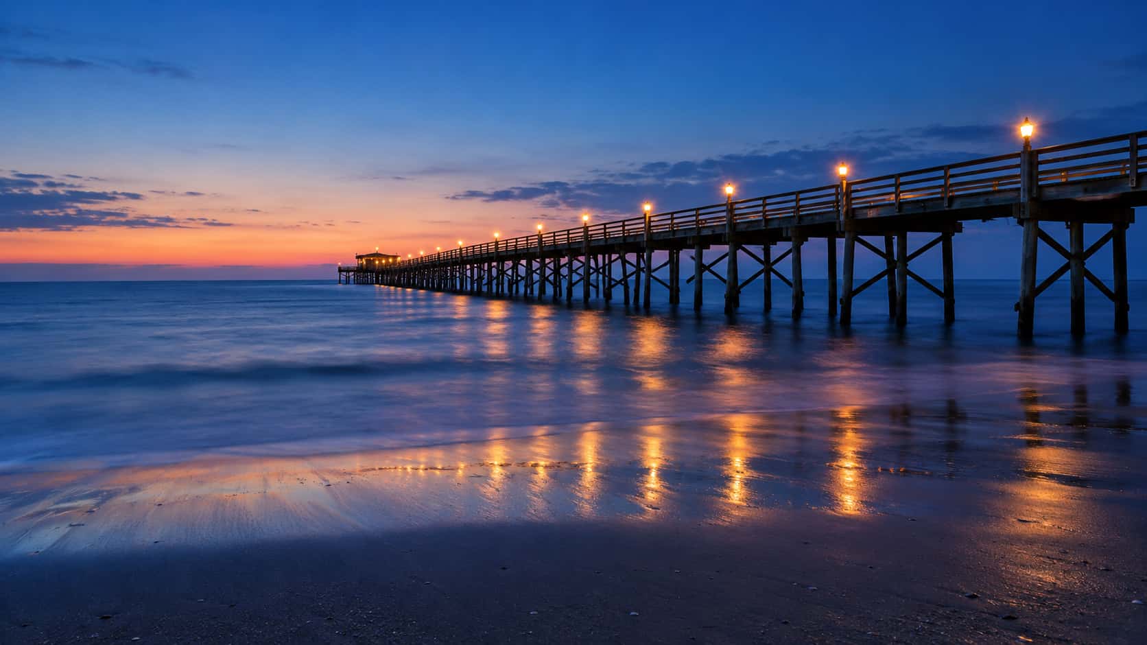 A long wooden pier with a string of warm amber lamps runs out over silky calm water from an empty sandy beach at blue hour.
