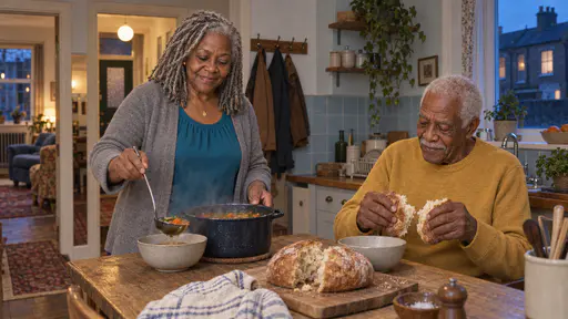 A silver-locked woman ladles steaming soup from a dark enamel pot into a bowl while a white-haired man tears a round loaf of bread at an oak kitchen table with pale blue tiles behind in warm dusk light.