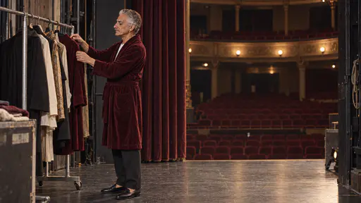A tall slim man in his early sixties with silver hair and one pearl earring in a plain burgundy robe adjusts plain costume garments on a rail at the edge of a small theatre stage, a half-drawn velvet curtain and empty seats beyond.
