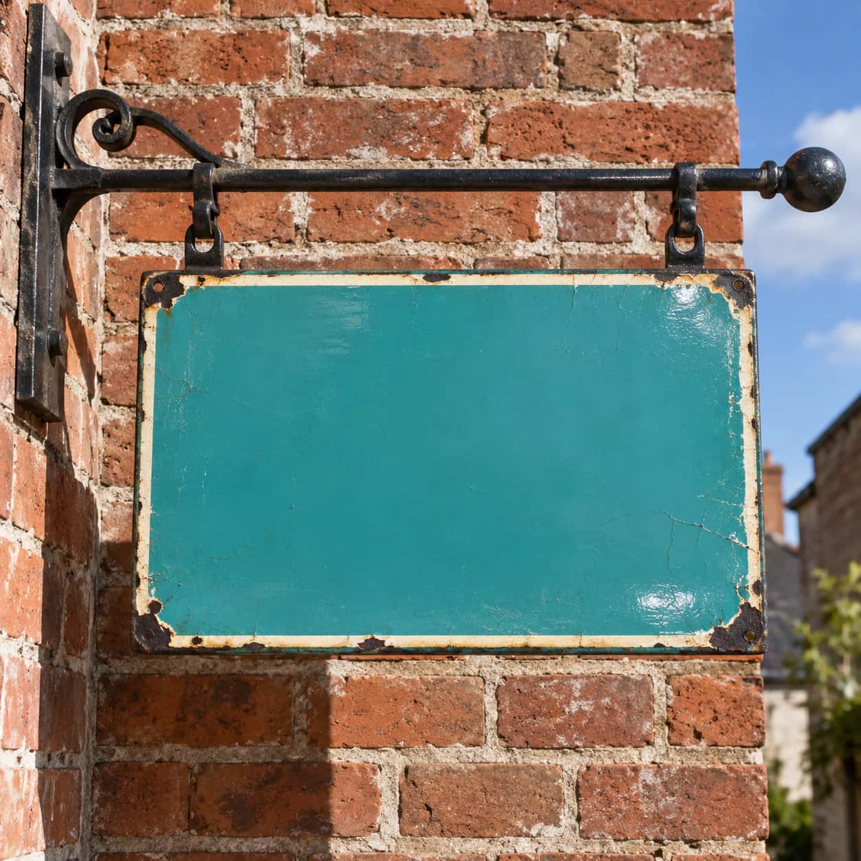 A blank turquoise enamel sign with chipped corners showing black iron and a worn cream border, hung on a scrolled iron bracket against weathered red brick, blue sky at the right, no lettering.