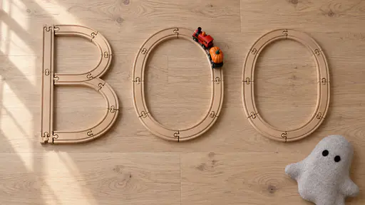 Top-down view of the word BOO made from curved and straight wooden toy railway track on a pale oak floor, a small red locomotive pulling an orange pumpkin wagon around the first O and a grey felt ghost cushion at the lower right.