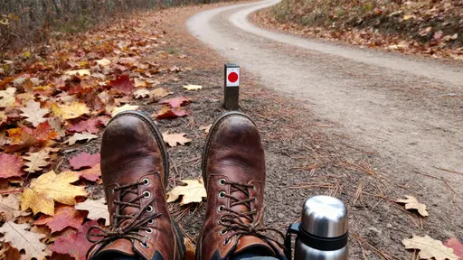 Worn leather boots pause on a leaf-strewn trail of russet maple and gold oak over compacted earth and pine needles, a stainless thermos cap nearby, soft overcast light balancing warm rusts and cool browns with shallow depth separating boots