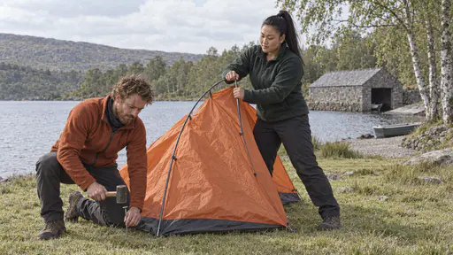 A lean bearded man in a rust fleece hammers a tent peg with a mallet while a broad-shouldered woman with a high ponytail holds the pole of a half-raised plain orange tent in a grassy clearing beside a lake in bright afternoon light.