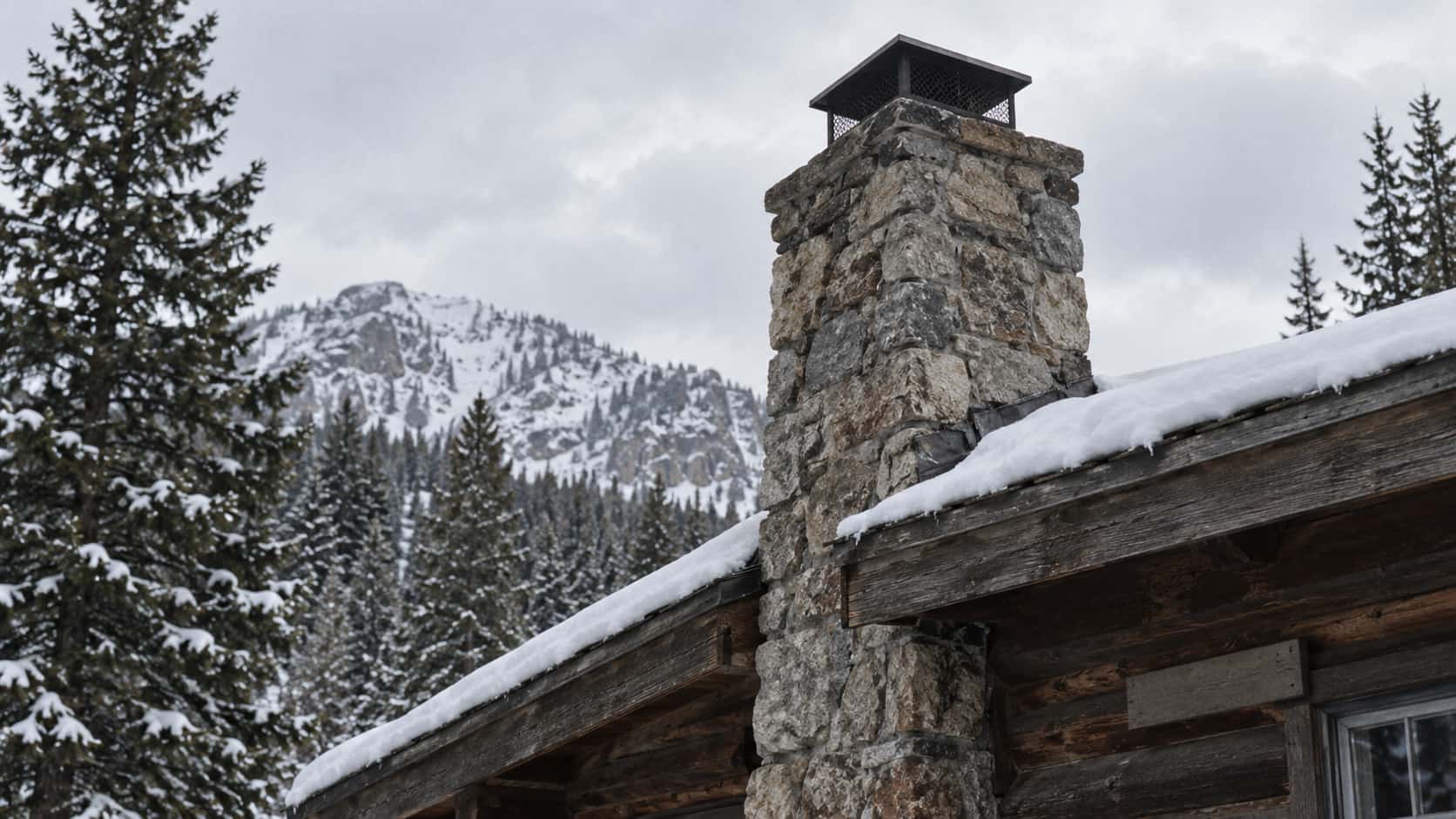 A stone chimney rises above the snow-lined roofline of a cabin with its cap clear of the snow in clear cold light.