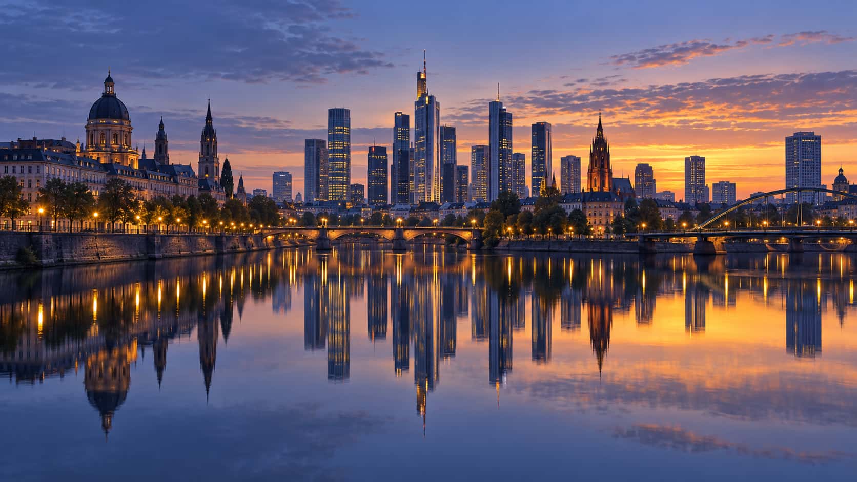A city skyline of glass towers and stone buildings stands across a calm river at first sunrise with peach light behind the roofs and a mirrored reflection.