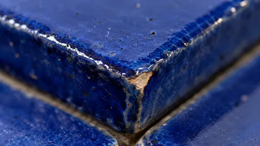 The corner of a handmade cobalt blue ceramic tile shows pooled glossy glaze, a tiny chip exposing pale clay and fine crazing in a razor-sharp macro.