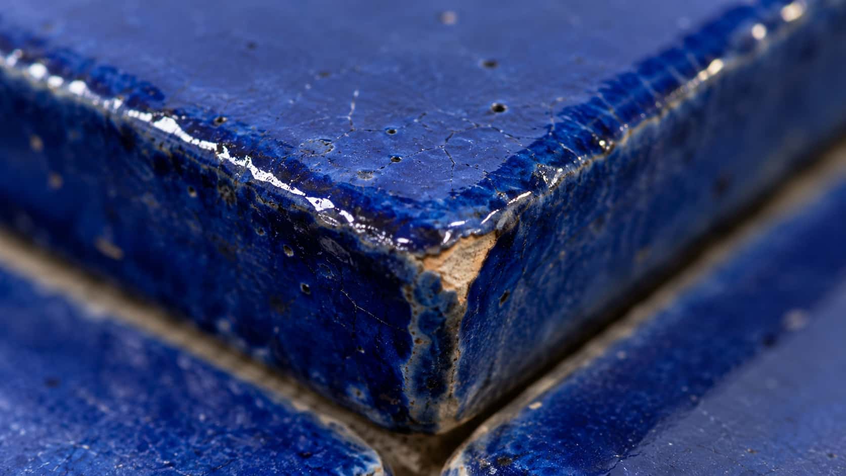 The corner of a handmade cobalt blue ceramic tile shows pooled glossy glaze, a tiny chip exposing pale clay and fine crazing in a razor-sharp macro.