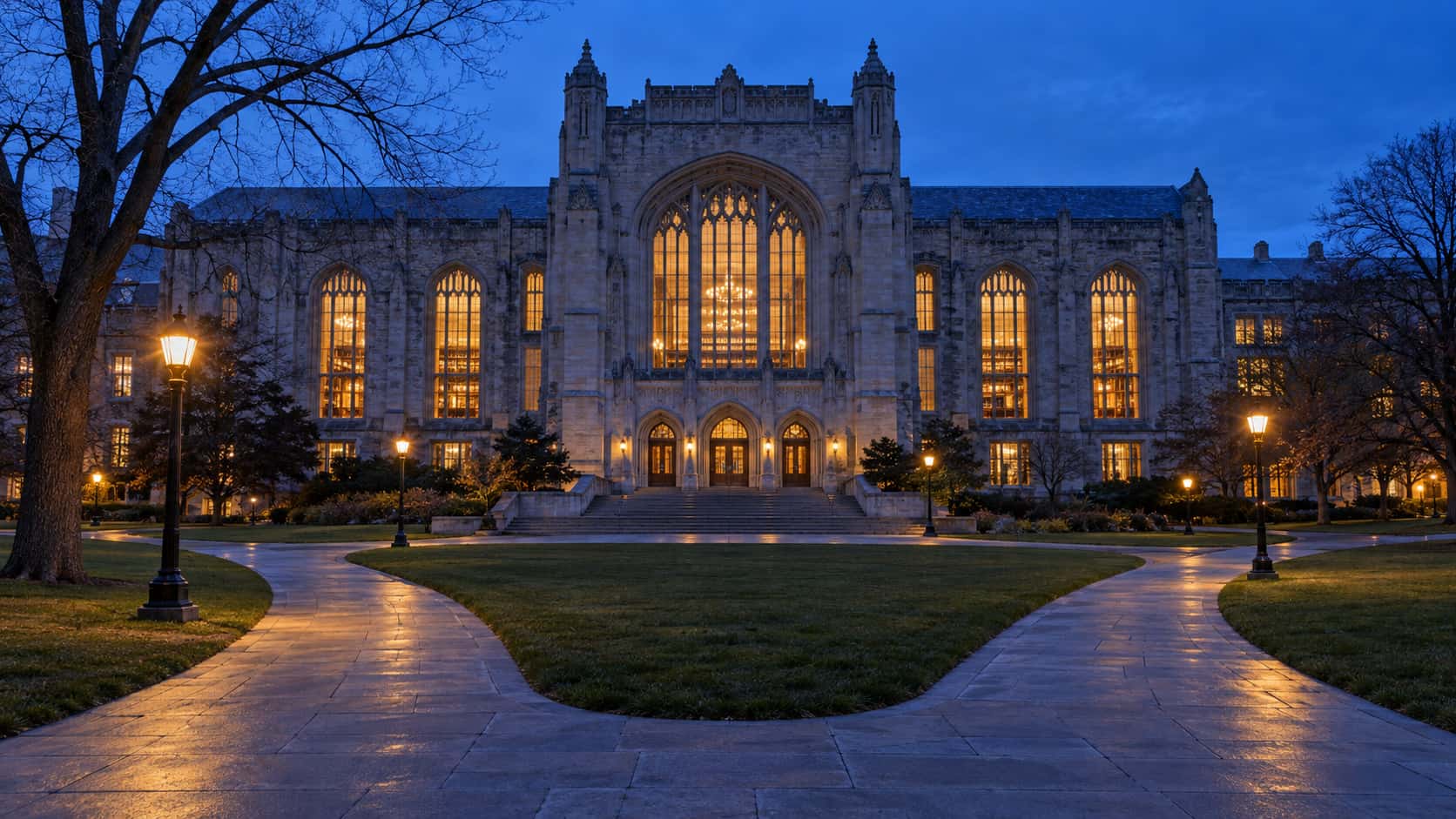 A grand stone college library with tall amber-lit arched windows stands across an empty quad at blue hour with curving paths leading to its stair.