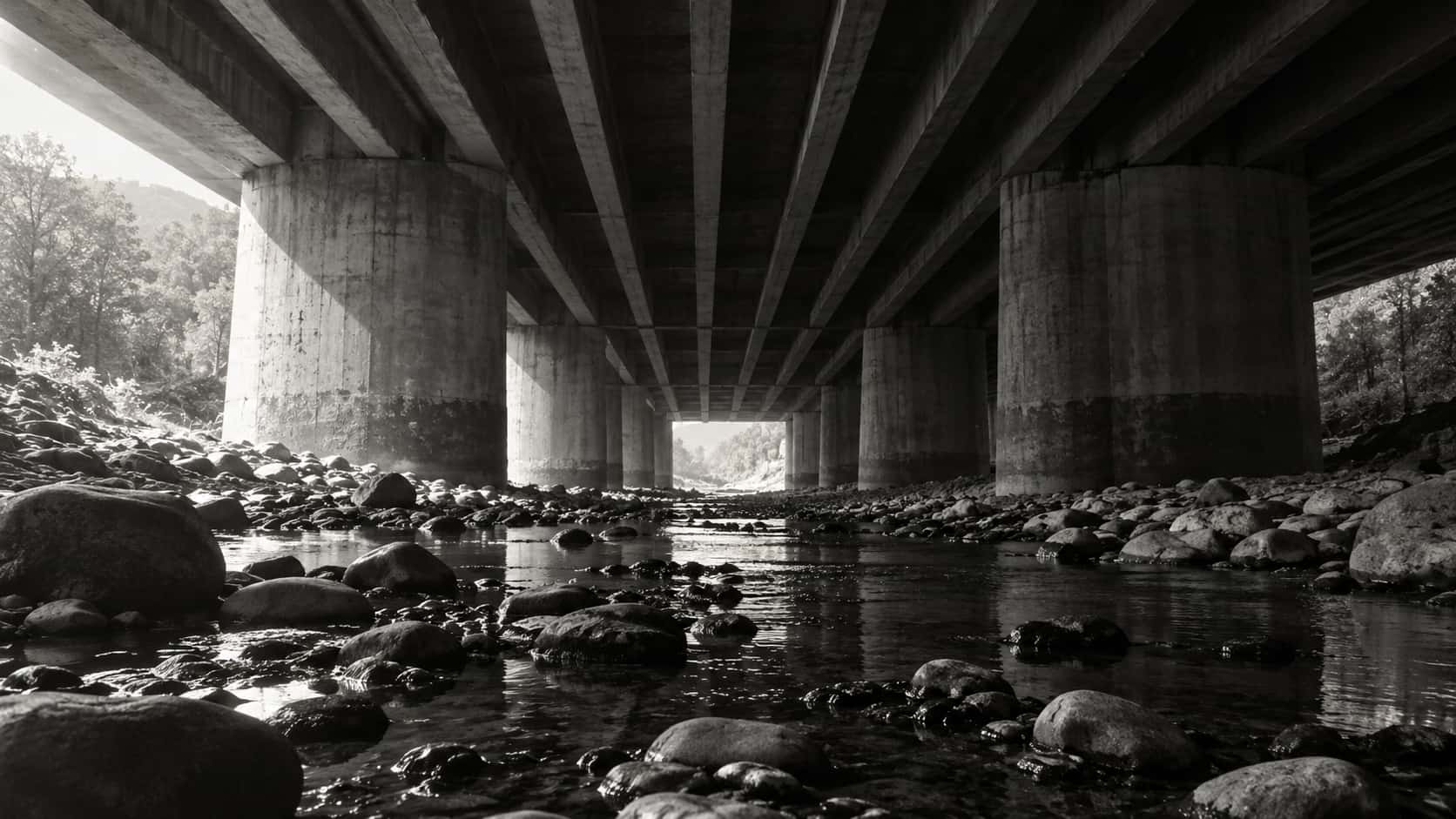 The underside of a concrete road bridge with massive piers and repeating ribbed beams seen from a dry riverbed with a thin stream and round stones.