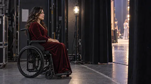 A woman with long dark wavy hair and a caramel streak in her plain black manual wheelchair and a long dark red velvet costume gown waits calmly in the wings beside a clear step-free side-stage route with her hands resting in her lap and her eyes toward the lit stage in cool ghost-light glow.