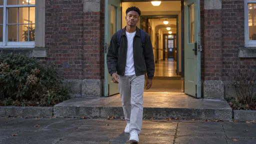 A tall young man with a coily twist-out in a charcoal jacket and navy rucksack walks easily out through a pale green double door toward the camera with a lit corridor behind him and warm light spilling onto the stone step at blue dusk.