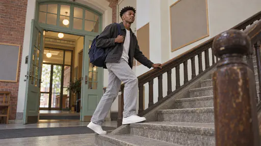 A tall young man with a coily twist-out in a charcoal jacket and navy rucksack climbs broad stone stairs two steps at a time with a hand trailing along a worn wooden banister in warm corridor light.