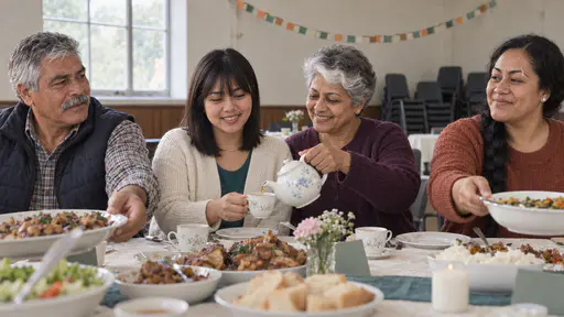 A young woman in a cream cardigan over a teal top sits at the head of a long table while an older woman in a plum cardigan pours tea and an older man in a navy gilet and a woman in a rust-orange jumper pass a dish of food in warm even hall light.