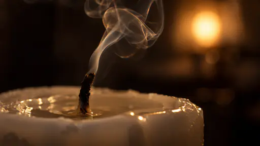 A thin wisp of smoke curls up from the blackened wick of a just-extinguished white candle in a razor-sharp macro with a clear wax pool.