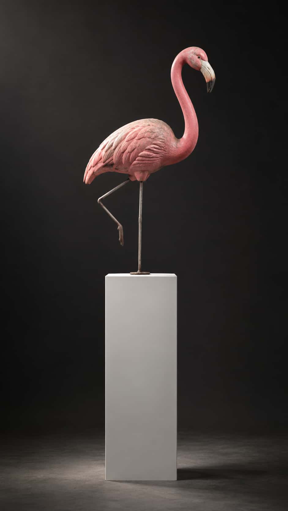 A sun faded pink plastic lawn flamingo with a bent leg stands on a tall white museum pedestal under formal light against a deep charcoal wall in a studio photograph.