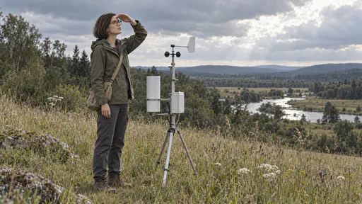A slim woman with a sleek dark bob and round tortoiseshell glasses in a waterproof olive jacket shades her eyes to look up at breaking clouds beside a small white weather station on a tripod with a slim rain gauge and a wind vane at the edge of a meadow with a stream glinting in the distance.