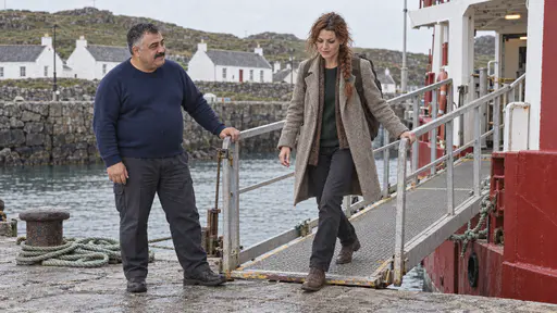 A heavyset man with a wide moustache in a navy sweater stands beside a gangway rail with a coiled rope at his feet while a freckled woman with an auburn braid and a rucksack steps down onto a grey stone landing below whitewashed island cottages.