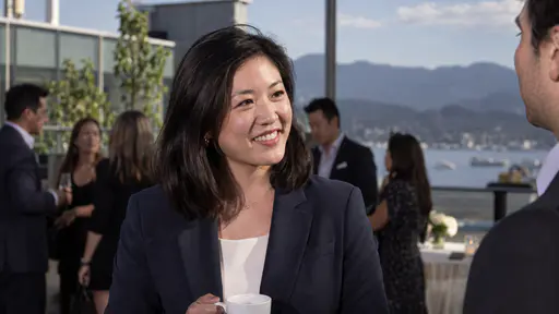 A fictional adult Chinese American founder, a woman with shoulder length black hair in a navy blazer, holds a plain coffee cup and smiles on a bright conference terrace in afternoon light.