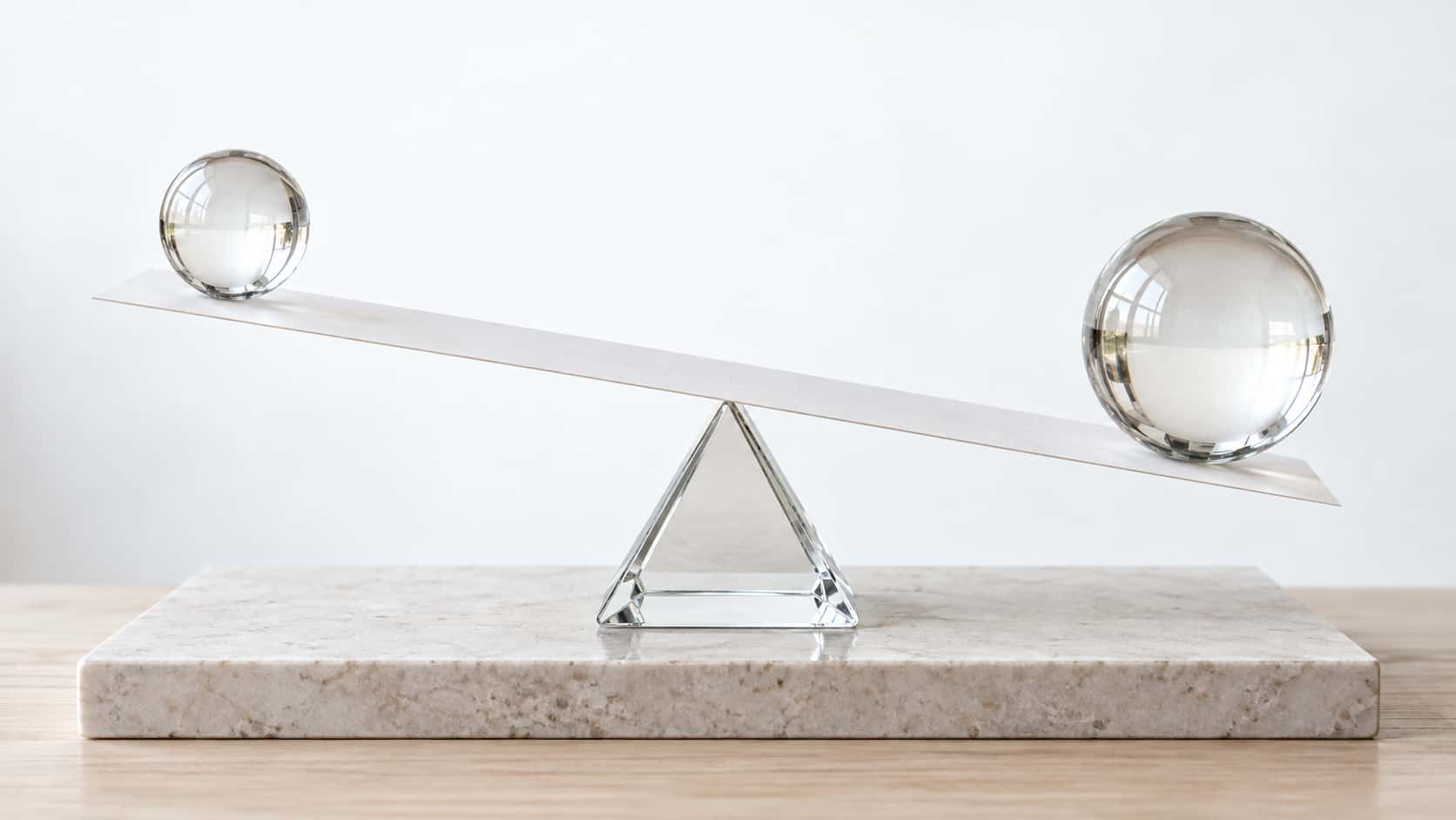 A long flat white paper beam rests across a single triangular glass fulcrum at its middle and carries a clear glass sphere on each end, the beam tilted gently toward the larger sphere, on a pale stone plinth in bright backlight.
