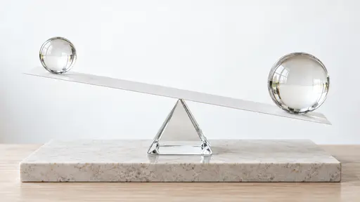A long flat white paper beam rests across a single triangular glass fulcrum at its middle and carries a clear glass sphere on each end, the beam tilted gently toward the larger sphere, on a pale stone plinth in bright backlight.