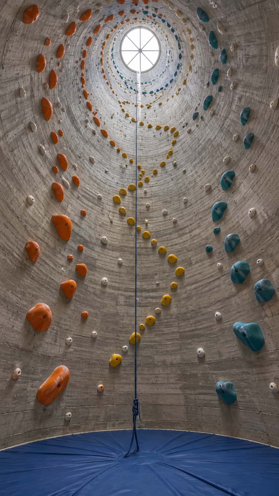 Looking straight up the inside of a round concrete silo turned climbing hall: coloured holds in orange, teal, yellow and white spiral up board-formed walls toward a circular skylight, a blue rope hangs to a round blue crash mat on the floor.
