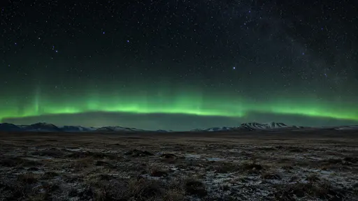 A low green aurora arc lies parallel to the horizon above a wide open plain with its lower edge sharply defined against the stars.